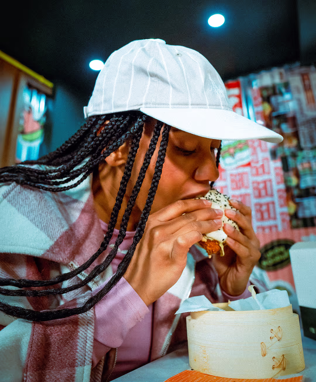 Une jeune femme, portant une casquette rayée rose et blanche, mange avec appétit un bao Krousty Sabaïdi au poulet dans un restaurant. Ses cheveux sont tressés et elle porte une veste à carreaux rose et blanche. Un panier vapeur en bois est visible devant elle.