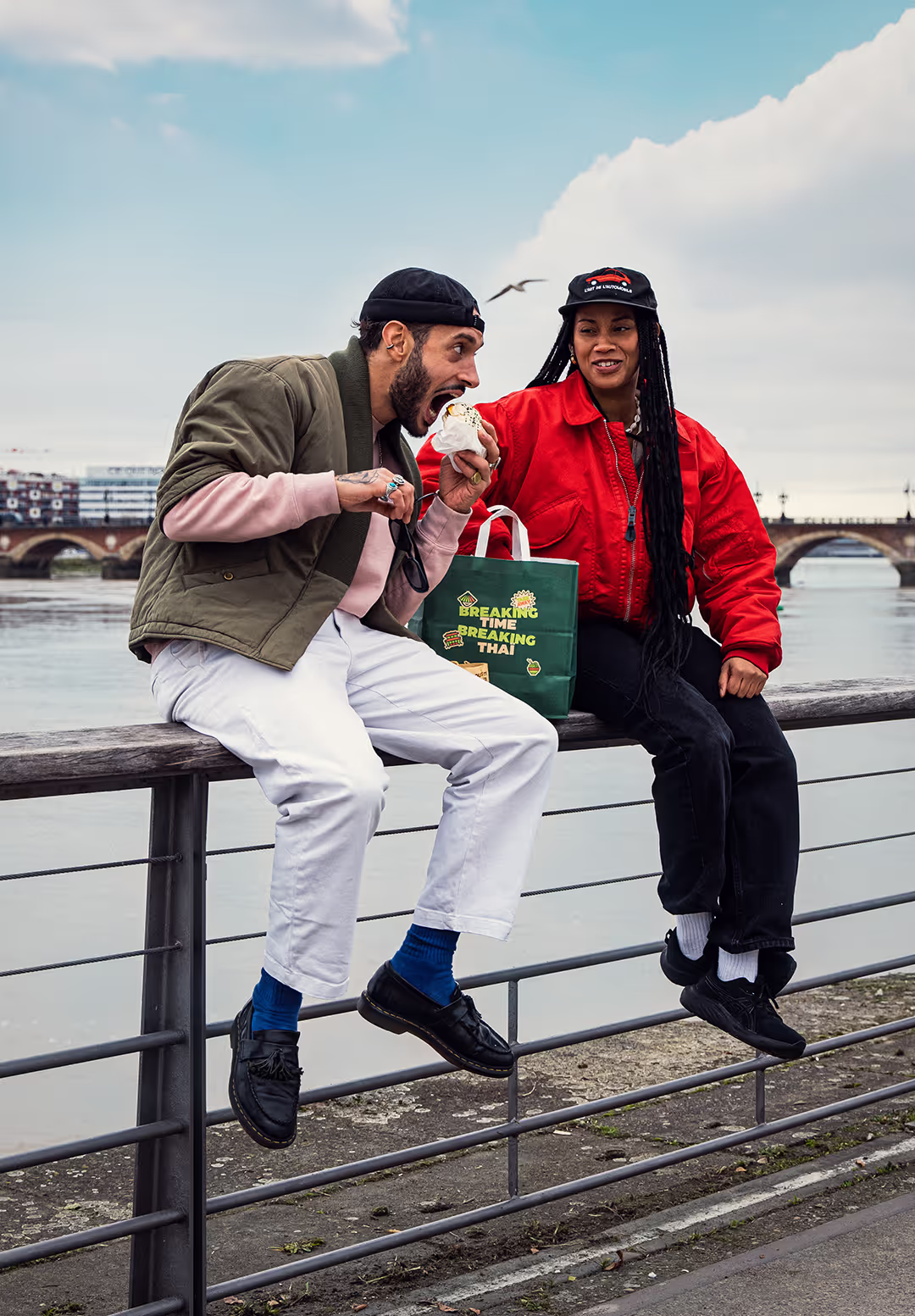 Un couple assis sur une barrière en bois au bord de la Garonne, avec le pont de pierre de Bordeaux en arrière-plan. L'homme porte une veste verte et blanche, et une casquette noire, et mange un bao en riant. La femme porte une veste rouge, une casquette noire et a de longues tresses. Elle regarde le bao de l'homme avec un grand sourire. Entre eux, il y a un sac de la marque Krousty Sabaïdi.