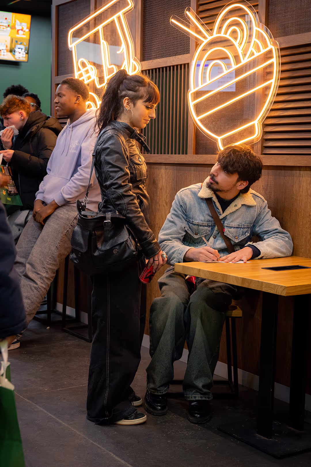 Deux jeunes gens discutent dans un restaurant thaï. Une femme se tient debout, penchée vers un homme assis à une petite table. La femme a les cheveux attachés en queue de cheval, porte une veste en cuir noir et un grand sac noir. L'homme assis, avec une barbe, est vêtu d'une veste en jean doublée de fourrure. Des néons en forme de bol de nouilles et d'autres dessins sont visibles sur le mur en bois.