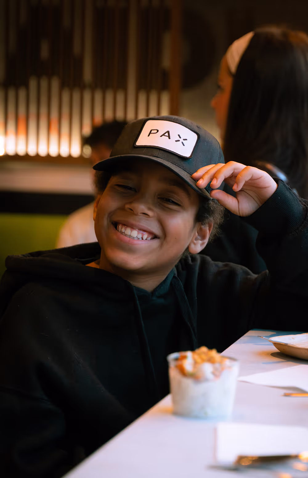 Jeune garçon souriant portant une casquette noire avec logo 'PAX' assis à une table de restaurant Krousty Sabaïdi avec de la nourriture.