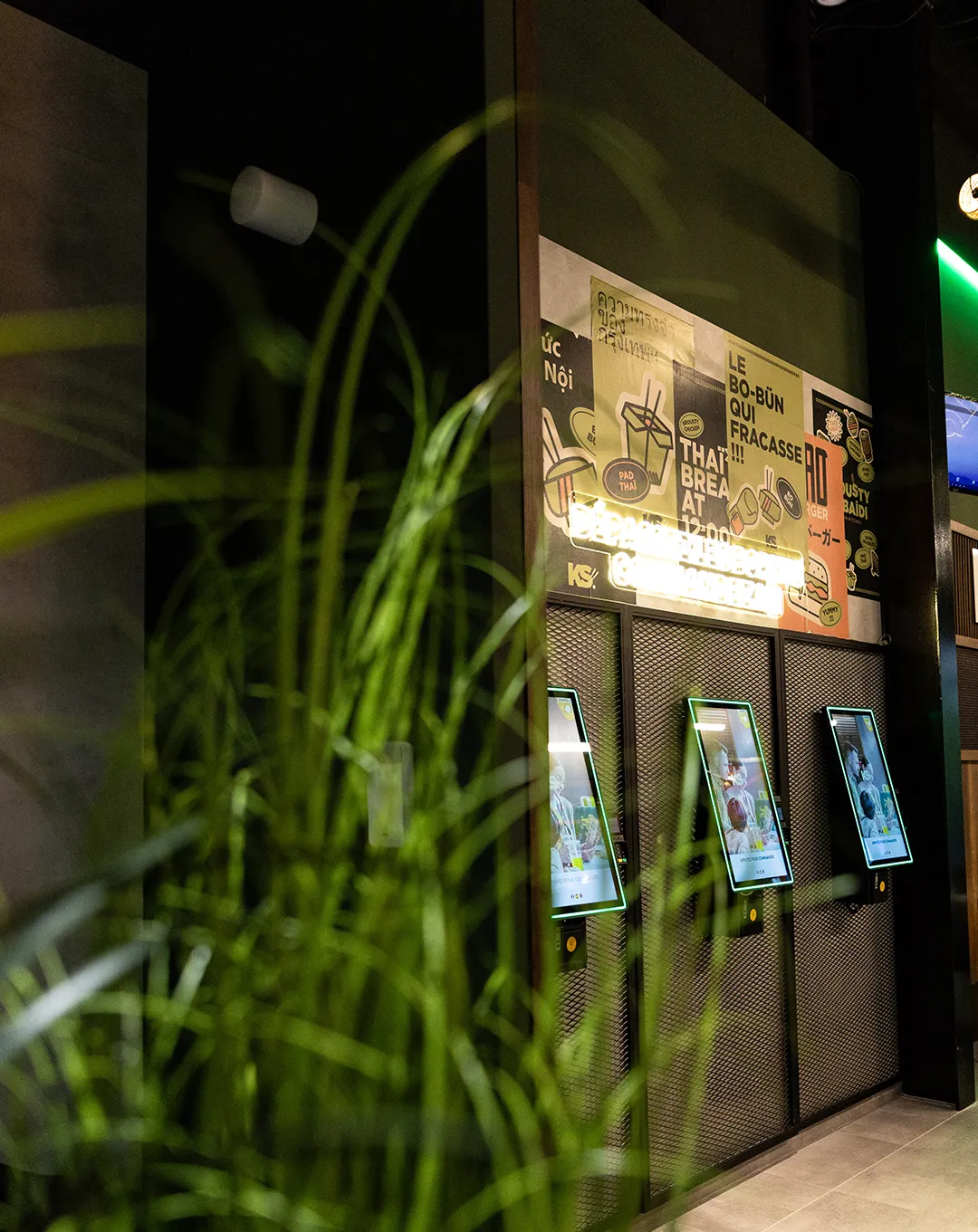 Intérieur de restaurant Krousty Sabaïdi avec bornes de commande tactiles, panneau publicitaire au plafond et plantes décoratives.