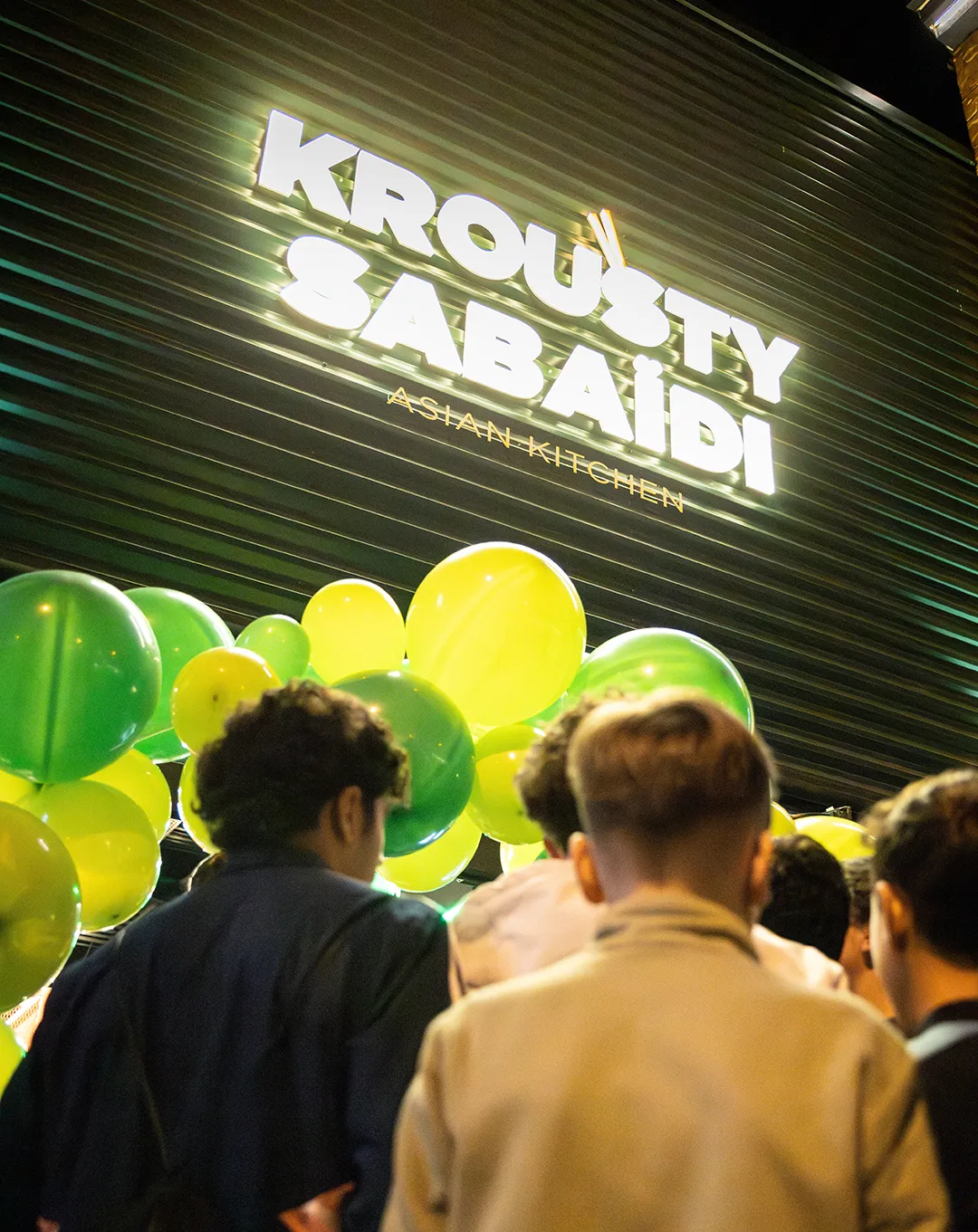 Enseigne lumineuse Krousty Sabaïdi Asian Kitchen la nuit avec foule et ballons verts et jaunes lors d'un événement d'ouverture.