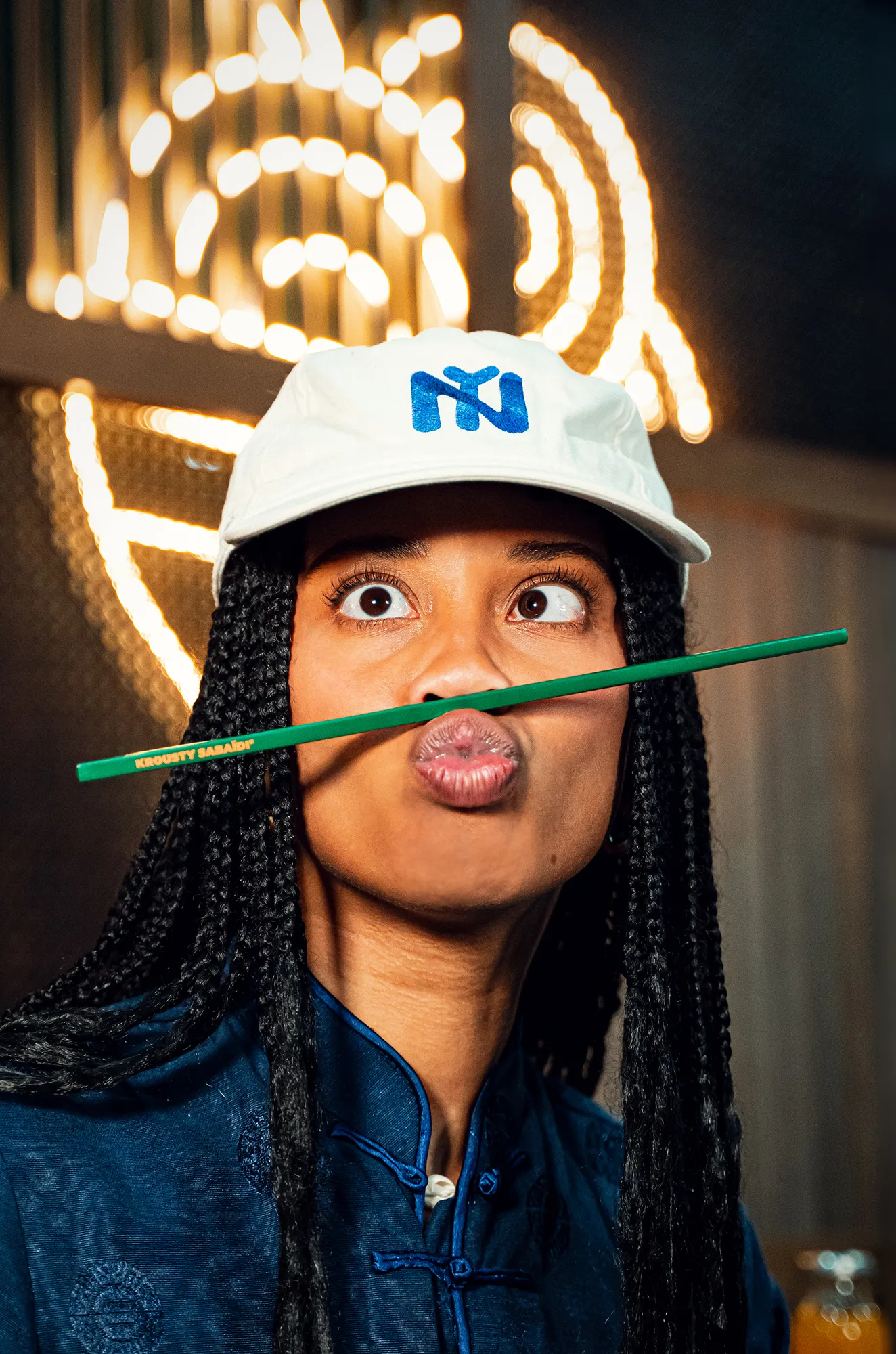 Femme souriante portant une casquette Krousty Sabaïdi, tenant une baguette verte de manière humoristique devant un décor lumineux.