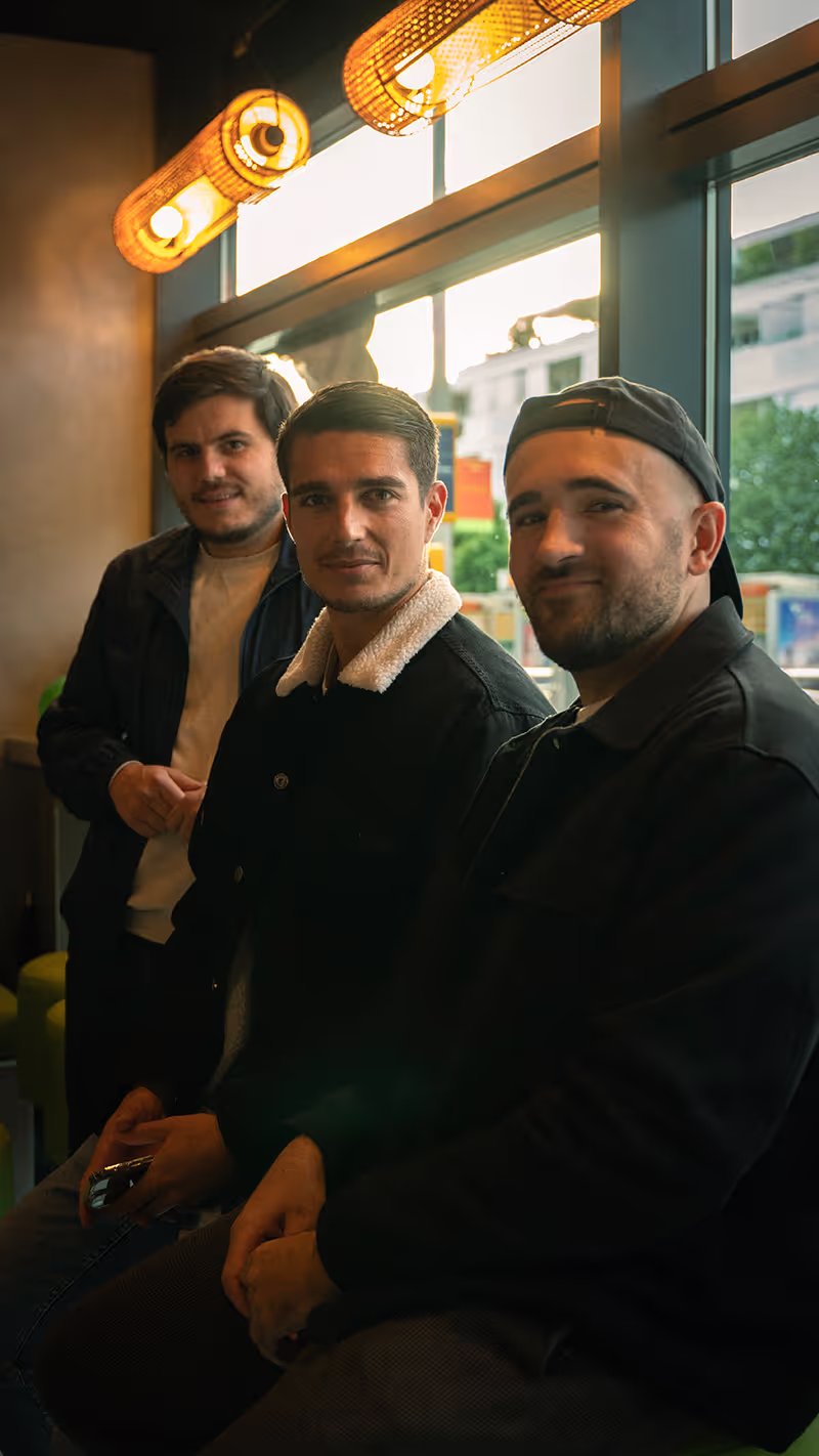 Trois hommes souriants posent ensemble dans un restaurant Krousty Sabaïdi, avec une lumière chaleureuse en arrière-plan. Photo d'amis en train de profiter d'un moment de détente dans l'établissement.