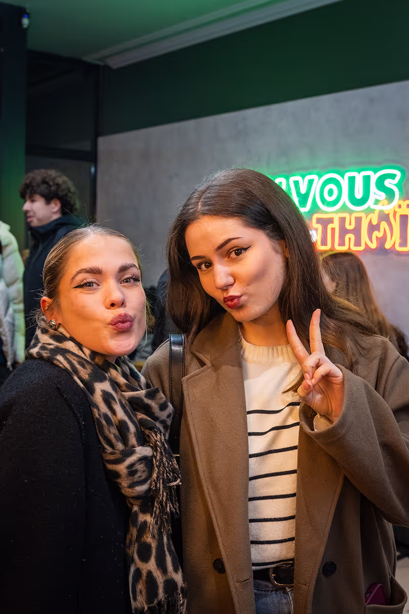 Deux jeunes femmes posant pour un selfie devant un restaurant Krousty Sabaïdi avec enseigne néon, l'une fait un bisou et l'autre un signe avec ses doigts.