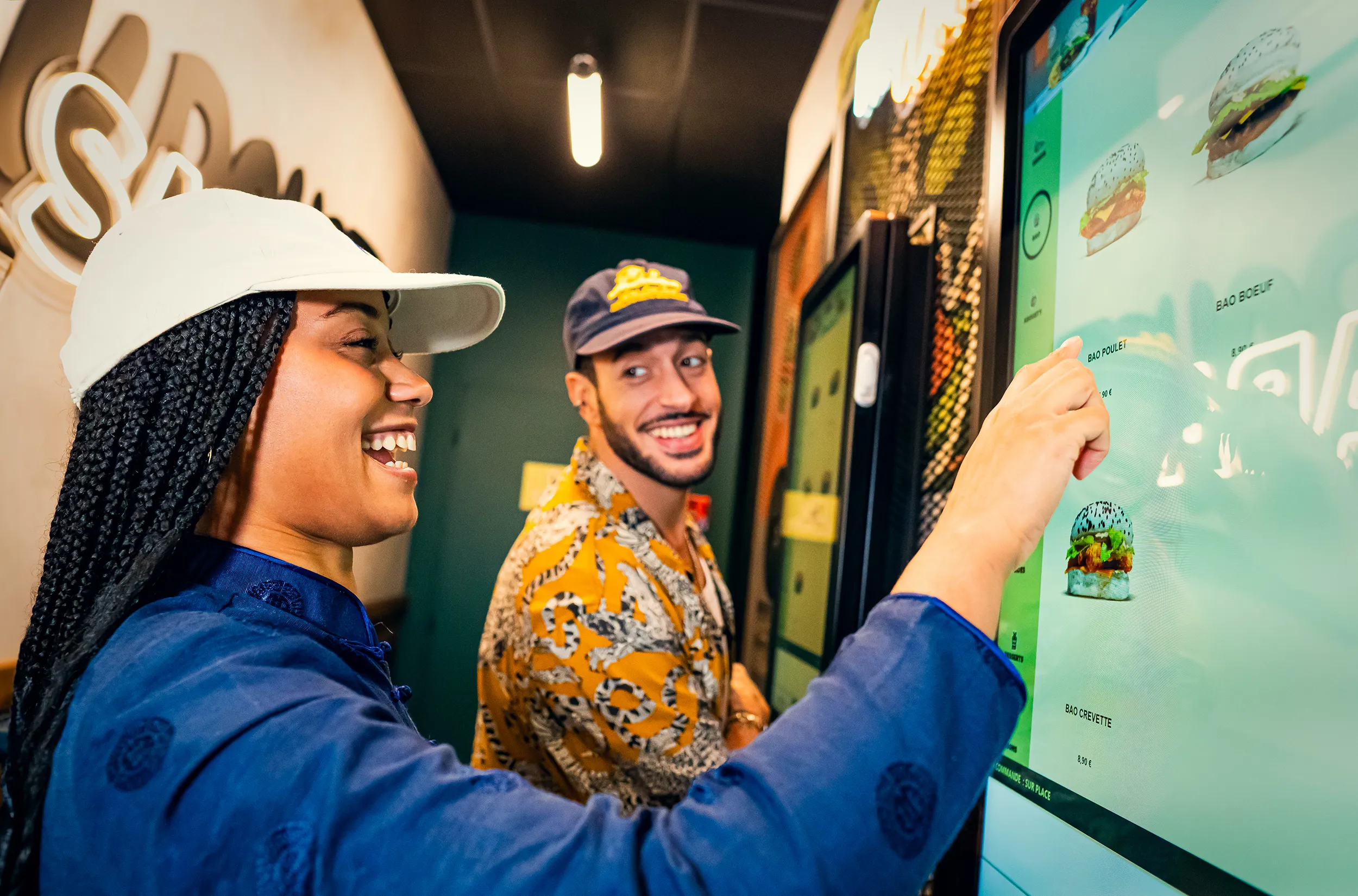 Deux clients souriants commandent sur une borne interactive dans un restaurant Krousty Sabaïdi. Une femme, portant une casquette blanche et une veste bleu foncé, touche l'écran. Un homme, coiffé d'une casquette bleue et vêtu d'une chemise à motifs jaune et noir, la regarde en souriant. L'écran de la borne affiche des images de baos.